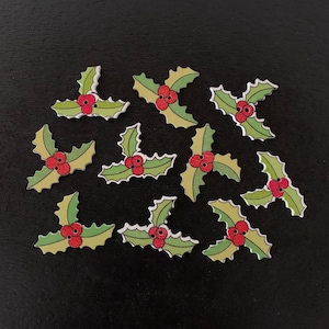 May include: A collection of holly leaf-shaped embellishments. Each wooden piece features green leaves with white trim and three red berries. These festive decorations are ideal for holiday crafts and seasonal projects.