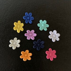 May include: Assortment of felt flower embellishments in various colors, including yellow, blue, pink, purple, white, orange, and teal. The flowers are arranged in a circular pattern on a black background.