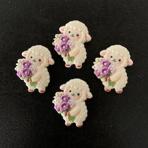 May include: Four cream-colored sheep figurines, each holding a bouquet of purple flowers. The sheep have pink accents on their hooves and ears. The background is a dark, textured surface.