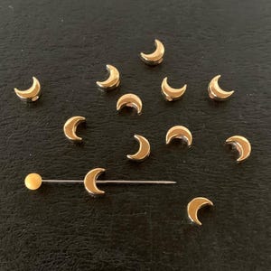 May include: Gold-colored crescent moon charms scattered on a dark surface. One charm is attached to a straight pin with a gold ball on the end. The charms are small and shiny, reflecting light.