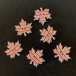 May include: Six wooden leaf-shaped buttons with red and orange striped patterns. Each button has two small holes for sewing. The buttons are arranged on a dark surface.