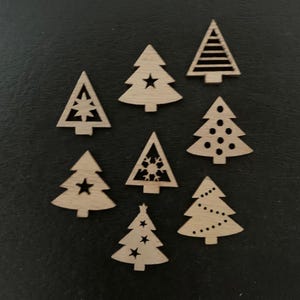 May include: A collection of eight small, wooden Christmas tree ornaments. Each tree has a unique design, including stars, dots, and geometric cutouts. The ornaments are light brown and arranged on a dark surface.