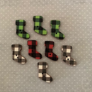 May include: A collection of eight wooden stocking-shaped buttons. The buttons feature a plaid pattern in green, red, and black and white. Each button has a small black button sewn on. The buttons are arranged on a light gray surface.