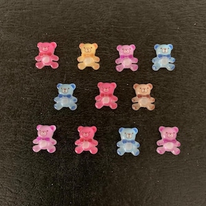 May include: A collection of twelve small, translucent teddy bear charms in various colors, including pink, yellow, blue, and purple. Each bear has a bow tie detail. The charms are arranged on a dark surface.