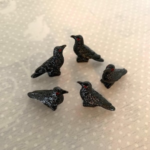 May include: Five small, black raven figurines with textured surfaces and red eyes are arranged on a light-colored, patterned surface. The birds are in various poses, some standing and some perched.