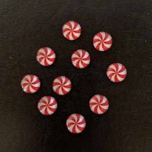May include: A collection of ten red and white peppermint candy-shaped embellishments. Each circular piece features a spiral design of red and white stripes. The candies are arranged on a dark surface.