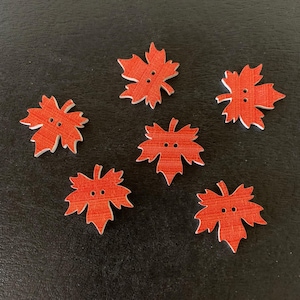 May include: Six wooden leaf-shaped buttons with a reddish-orange finish and two small holes. The buttons have a white outline and are arranged on a dark surface. These buttons are ideal for sewing and craft projects.
