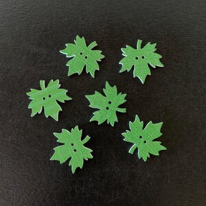 May include: Six green leaf-shaped wooden buttons with two holes each. The buttons have a textured appearance and a white outline. The buttons are scattered on a dark surface.