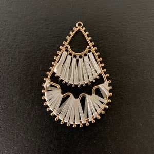 May include: A gold-colored, teardrop-shaped earring pendant with white thread detailing. The pendant has a delicate, openwork design with a small loop at the top for hanging. The white thread is arranged in a fringe pattern.
