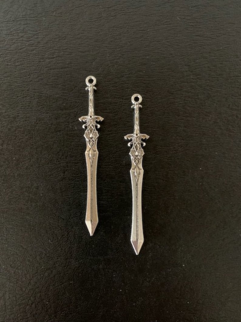 Pu&ograve; includere: Due ciondoli a forma di spada color argento con intricati disegni dell'elsa e lame appuntite. Ogni ciondolo ha un piccolo anello nella parte superiore per il fissaggio a gioielli o altri lavori artigianali. Sfondo scuro.