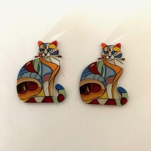 May include: A pair of colorful cat earrings with a stained glass design. The cats are sitting with their tails curled around their legs. The earrings are made of metal and have a shiny finish.