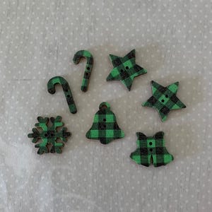 May include: A collection of seven wooden buttons featuring a green and black buffalo plaid pattern. The set includes two candy canes, two stars, one snowflake, one bell, and one bow shape, each with two small holes for sewing or crafting purposes.