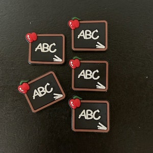May include: Five small, square, black chalkboard charms with the letters "ABC" in white, a white arrow, and a red apple. The charms have a brown border and are scattered on a dark surface. The charms are likely for shoes or accessories.