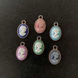 May include: A collection of oval cameo pendants in various colors, including blue, pink, teal, purple, green, and gray. Each pendant features a white profile and a gold-colored frame with a beaded edge. They have a small loop at the top for attaching to jewelry.