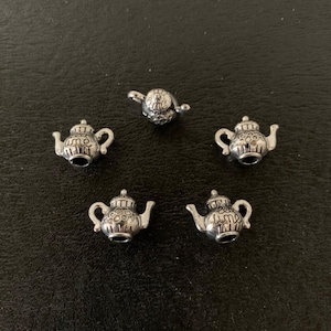 May include: Five small, silver-colored teapot charms. Each charm has a detailed design, including a spout, handle, and lid. The teapots have the word "time" on the front. The charms are arranged on a dark surface.