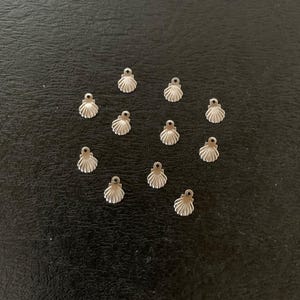 May include: A collection of 14 silver-toned metal seashell charms, arranged in a heart shape on a black surface.