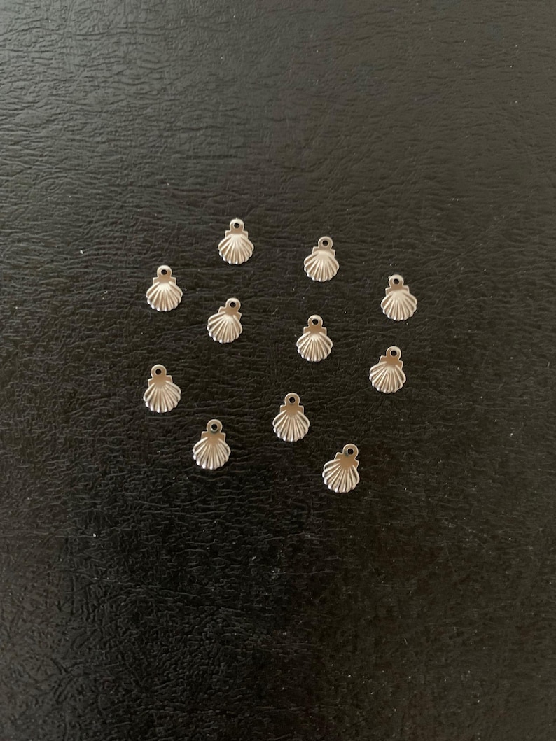 Puede incluir: Una colecci&oacute;n de 14 abalorios de concha de mar de metal plateado, dispuestos en forma de coraz&oacute;n sobre una superficie negra.