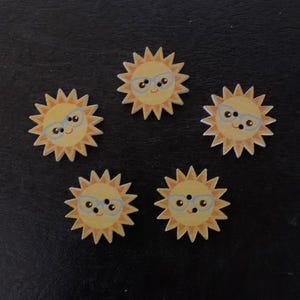 May include: Five wooden buttons shaped like smiling suns with yellow rays, wearing blue glasses. The suns have a light yellow face and are arranged in a circular pattern on a dark surface. Each button has two small holes for sewing.