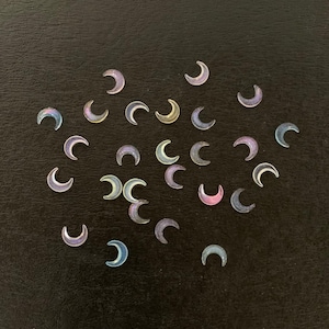 May include: A scattering of iridescent crescent moon shaped stickers on a black surface. The stickers are various shades of blue, purple, and pink.