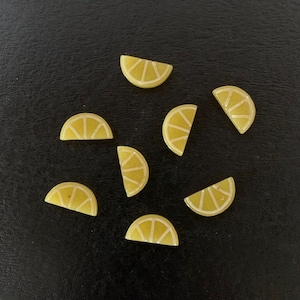 May include: Seven yellow lemon slice-shaped decorative objects with white detailing are scattered on a dark surface. The objects are arranged in a circular pattern, each with a curved shape and a textured appearance, resembling miniature citrus fruit.