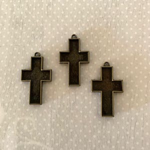 May include: Three bronze-colored cross pendants with a dark fill, each featuring a small loop at the top for hanging. The crosses are displayed on a white surface with a subtle polka dot pattern.