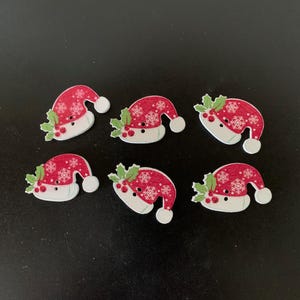 May include: Six flat buttons shaped like Santa hats. Each hat is red with white snowflake patterns, a white brim, a white pom-pom, and green holly leaves with red berries. Each button features two small holes in the center for attachment purposes.