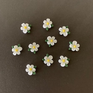 May include: Ten white daisy flower embellishments with yellow centers and green leaves. The flowers are arranged in a circle on a dark gray background.