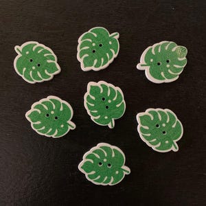 May include: Seven green leaf-shaped wooden buttons with white edges and two holes each. The buttons are arranged on a dark surface. The buttons are a tropical theme.