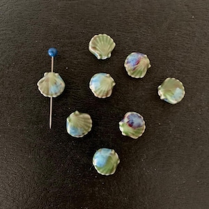 May include: A collection of small, ceramic seashell beads in shades of green, blue, and white. One bead is attached to a pin with a blue head. The beads are arranged on a dark surface.