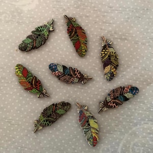 May include: A collection of seven decorative feather-shaped buttons. Each button features a unique pattern with a mix of colors including green, red, yellow, and blue. The buttons are made of wood and have a metallic silver button shank.