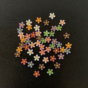 May include: A collection of small, colorful flower-shaped embellishments. The flowers are in various colors, including white, pink, orange, yellow, blue, and brown. They are scattered on a dark, textured surface.