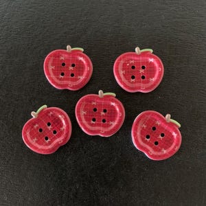 May include: Five red apple-shaped buttons with a plaid pattern. Each button has a green stem and four holes for sewing. The buttons are arranged on a dark surface.