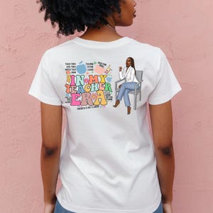 Pu&ograve; includere: Maglietta bianca con un design grafico colorato che presenta una donna seduta su una sedia. La grafica include il testo "In My Teacher Era" con uno schema di colori arcobaleno e altro testo che include "Teaching Future Leaders", "Read More Books" e "Progress is not a Linear".