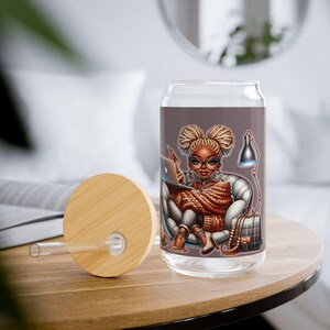 May include: A glass tumbler with a brown background and a cartoon illustration of a woman with brown skin and braids wearing glasses and a brown sweater, sitting in a chair with a laptop and a lamp. The tumbler has a bamboo lid and a clear straw.