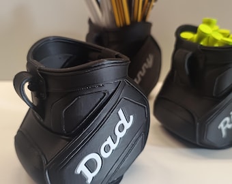 Könnte beinhalten: Schwarze Schreibtisch-Organizer in Golfbag-Form mit weißer Schrift. Einer trägt die Aufschrift "Dad", ein anderer "Ricky" und ein dritter "Sunny". Die Taschen sind mit Pinseln und Golf-Tees gefüllt. Ein einzigartiges Geschenk.