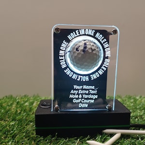 以下が含まれることがあります： ゴルフボールのデザインと「HOLE IN ONE」という文字がボールの周りに繰り返されている透明なアクリル製のプレート。プレートは、側面にスイッチが付いた黒いベースの上に置かれています。「Your Name」、「Any Extra Text」、「Hole & Yardage」、「Golf Course」、「Date」というテキストは、ゴルフボールの下にあります。