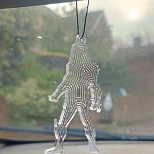 May include: Clear acrylic Bigfoot car charm. The charm is a silhouette of the legendary creature, with a detailed, layered design. It hangs from a black cord, perfect for rearview mirror decoration. The background is a blurred view of a sunny outdoor scene.
