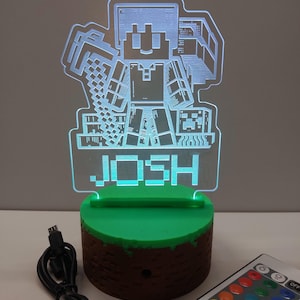 May include: A Minecraft-themed LED lamp featuring a pixelated character holding a pickaxe. The lamp has a green base and the name "JOSH" is illuminated. A remote control and USB cable are also visible.