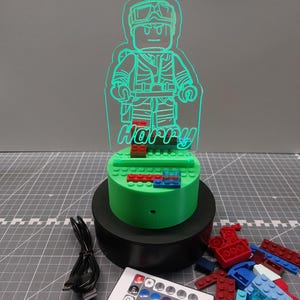 May include: A green LED night light in the shape of a Lego-style figure, with the name "Harry". The light is on a green base with red and blue blocks, on a black stand. A remote control and USB cable are also included.