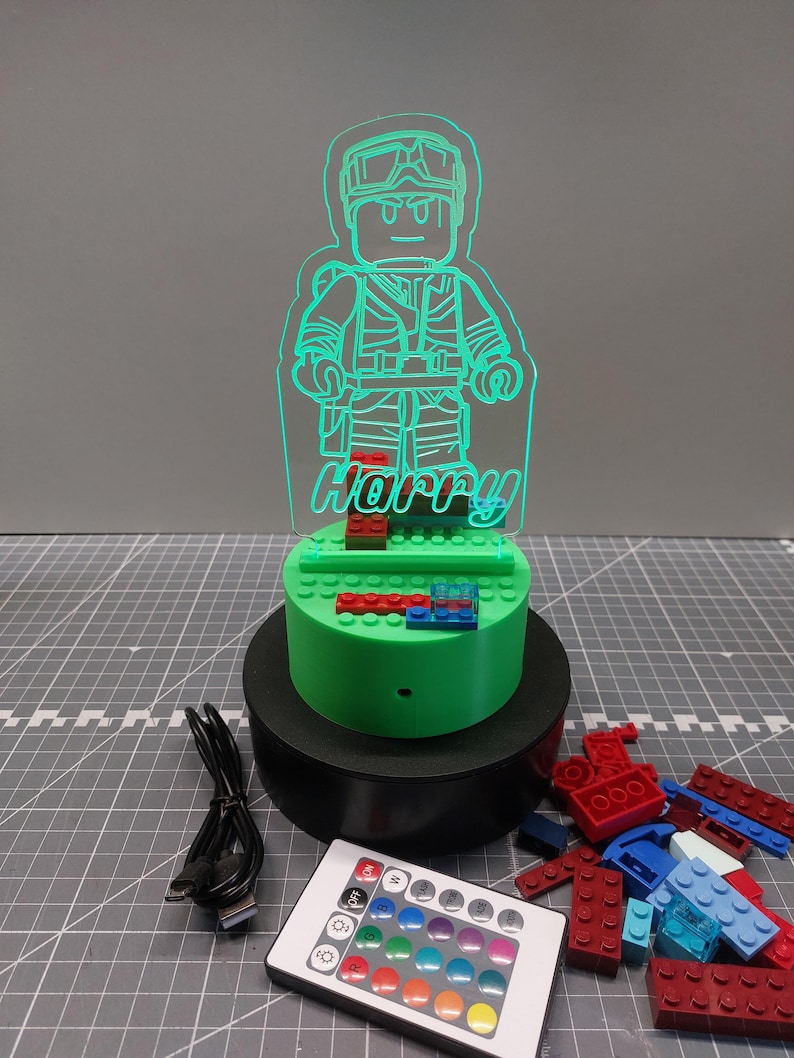 May include: A green LED night light in the shape of a Lego-style figure, with the name "Harry". The light is on a green base with red and blue blocks, on a black stand. A remote control and USB cable are also included.