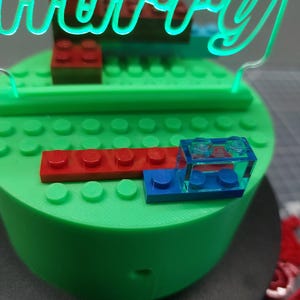 May include: A green, round cake base with red and blue toy blocks. A neon green sign that says "Harry" is on top of the cake. The cake is decorated with toy blocks.