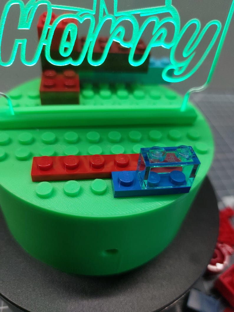 May include: A green, round cake base with red and blue toy blocks. A neon green sign that says "Harry" is on top of the cake. The cake is decorated with toy blocks.