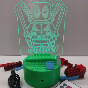 May include: A green LED night light with a Deadpool design. The light has a clear acrylic panel with a Deadpool figure and the name "Daniel" etched into it. The light sits on a green base with Lego-style studs and includes a remote control.