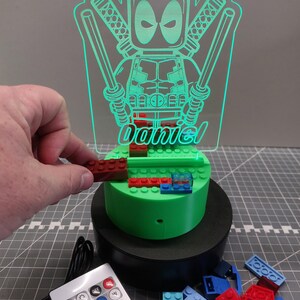 May include: A green LED night light with a Deadpool design and the name "Daniel". The light sits on a base with building blocks and includes a remote control. The light is powered by a USB cable.