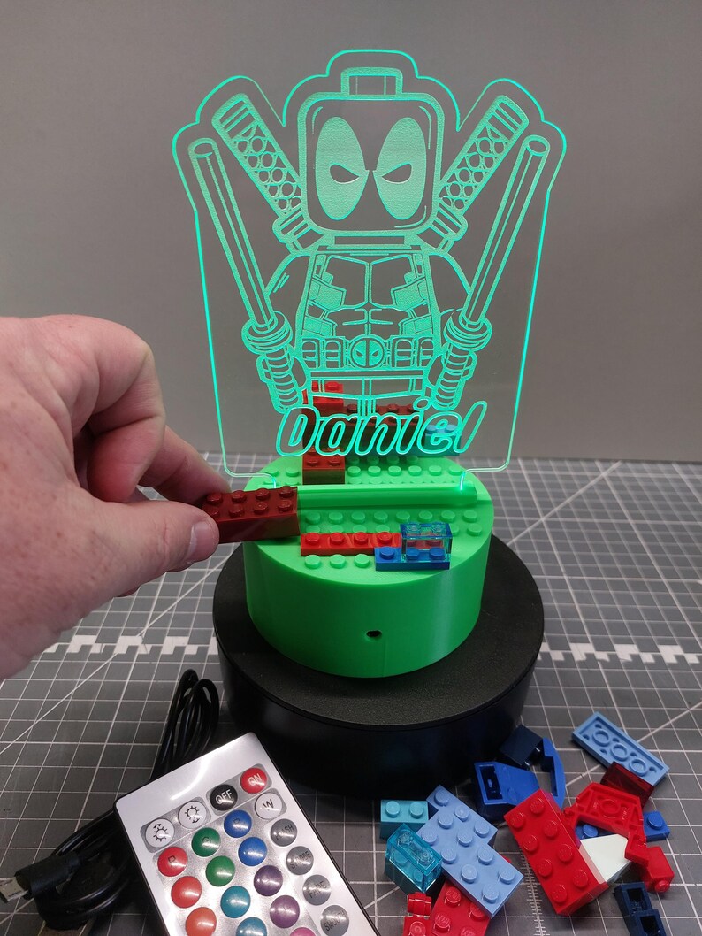 May include: A green LED night light with a Deadpool design and the name "Daniel". The light sits on a base with building blocks and includes a remote control. The light is powered by a USB cable.