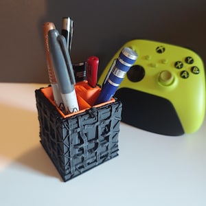 Pode incluir: Um porta-canetas geométrico preto e laranja cheio de canetas e marcadores. Um controle Xbox verde limão está no fundo. O porta-canetas tem um design texturizado em forma de cubo.