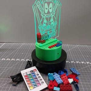 May include: A green Deadpool-themed LED night light with the name "Daniel" etched into the acrylic. The light sits on a green base with a black rotating stand. A remote control and colourful building blocks are also present.