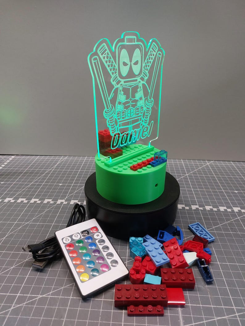 May include: A green Deadpool-themed LED night light with the name "Daniel" etched into the acrylic. The light sits on a green base with a black rotating stand. A remote control and colourful building blocks are also present.
