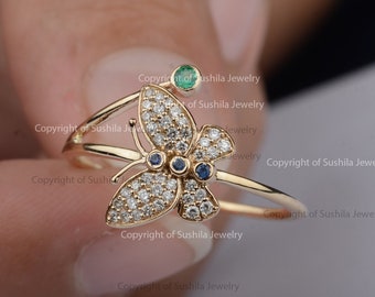 May include: A gold butterfly ring with a green gemstone on the top and two blue gemstones on the butterfly's wings. The butterfly is covered in small white gemstones.