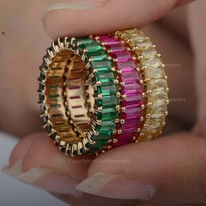 May include: Three gold rings with emerald green, pink, and yellow baguette-cut gemstones. The rings are stacked on top of each other.